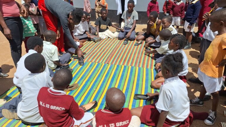 Children sit in a colorful circle on a mat, playing 'This Is Not My Shoe!' at All Is Grace School.