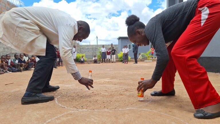 Two teachers at All Is Grace School laugh and race during a Fun Friday game involving plastic bottles, with children cheering around them.