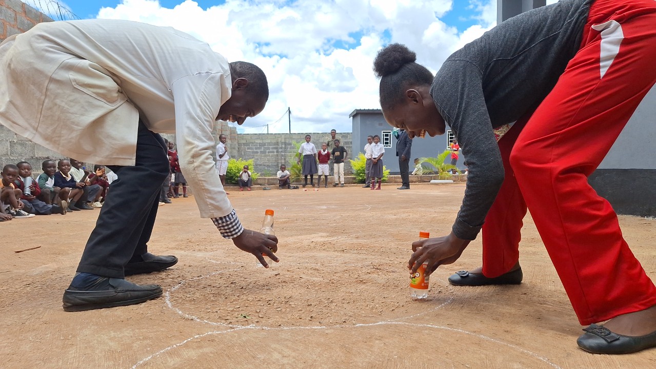 Two teachers at All Is Grace School laugh and race during a Fun Friday game involving plastic bottles, with children cheering around them.