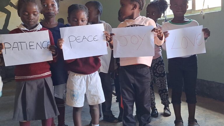 Children at All Is Grace School hold signs reading Patience, Peace, Joy, and Love as they present on the Gifts of the Holy Spirit.