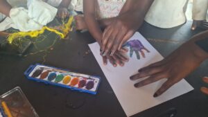 A teacher guides a child’s hand onto watercolor-painted paper to create a colorful handprint during an art lesson at All Is Grace School.