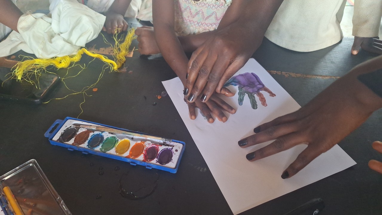 A teacher guides a child’s hand onto watercolor-painted paper to create a colorful handprint during an art lesson at All Is Grace School.