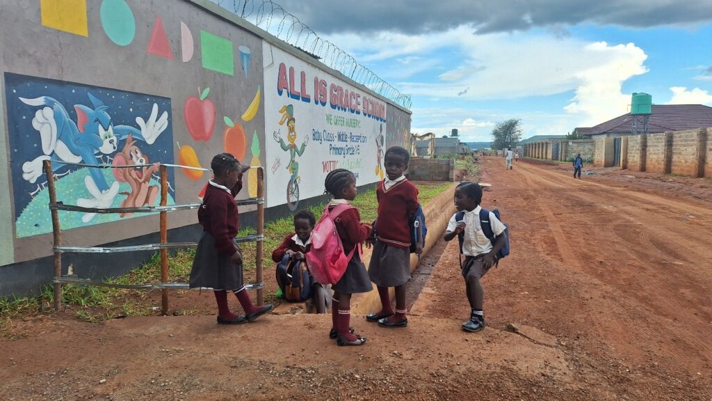 Children in school uniforms stand and talk beside a painted wall outside All Is Grace School along a dirt road.