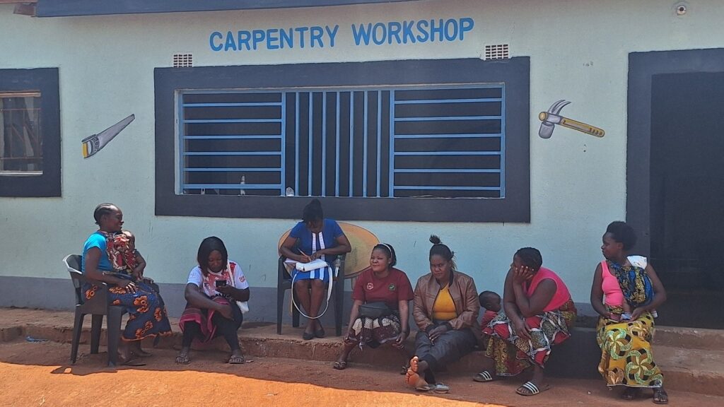 Women sit together outside a building labeled “Carpentry Workshop,” some holding young children.