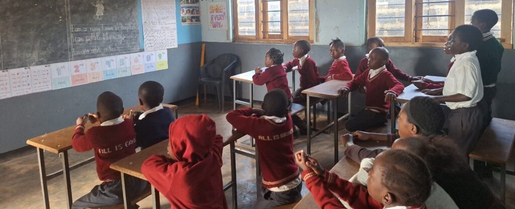 Students in school uniforms sit at desks inside a classroom, facing the front during a lesson.