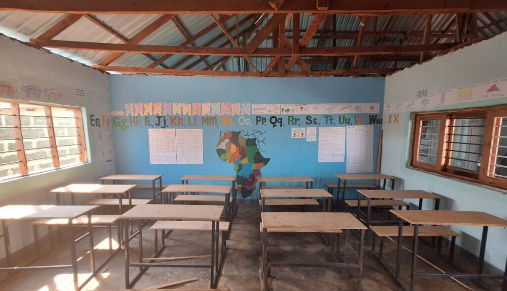 An empty primary school classroom with wooden desks, painted alphabet letters, and learning posters on the walls.
